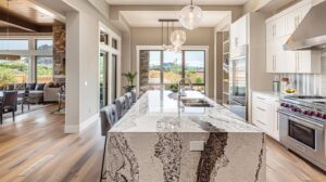 Luxurious modern kitchen featuring stunning natural stone countertops, large granite or quartz island with rich veining patterns in warm gray and white tones, polished surface with deep luster reflecting soft natural daylight from oversized windows, contemporary white shaker cabinets with brushed nickel hardware, professional stainless steel appliances, pendant lighting above island, open-concept design with comfortable seating area visible in background, hardwood or LVP flooring, fresh flowers or fruit bowl on countertop, inviting warm atmosphere, late morning golden sunlight streaming through windows creating soft shadows, professional architectural photography, impeccable stone fabrication quality, pristine condition showcasing natural stone beauty and durability, crystal clear details, elegant and functional kitchen space, no text, no people whatsoever