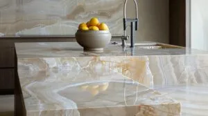 A high-end, professional architectural shot of a luxurious kitchen island featuring a thick Taj Mahal Quartzite countertop with soft, flowing cream and grey veins. On the surface, a minimalist ceramic bowl with lemons and a high-quality stainless steel faucet are visible. The stone has a polished finish that reflects a warm, bright kitchen interior. The lighting is soft and natural, emphasizing the depth and unique patterns of the natural stone. No people, no text, 8k resolution, architectural magazine style, sharp focus on the stone's mineral details.